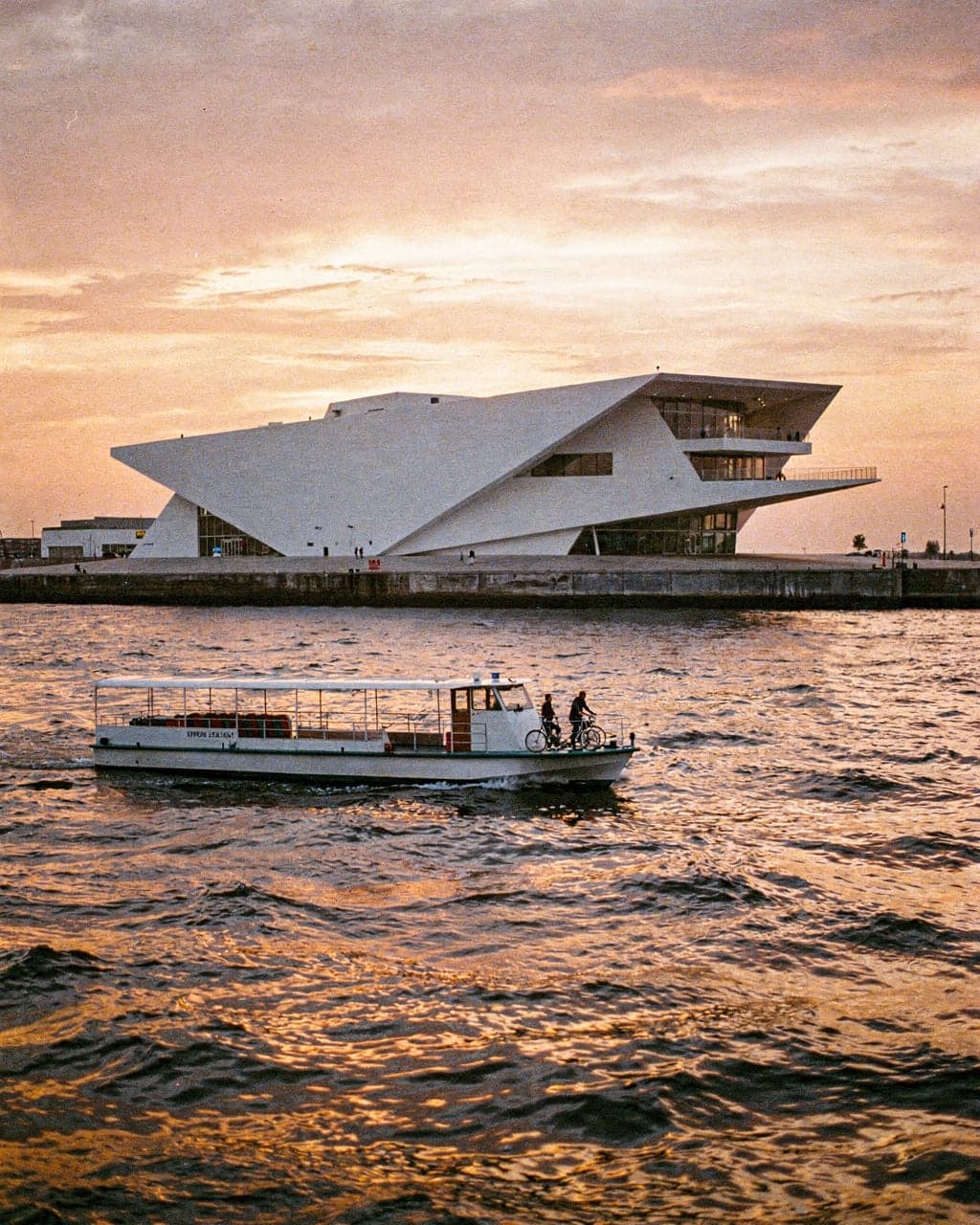 The white, angular EYE Filmmuseum on the north bank of the IJ at sunset, with a ferry crossing the water