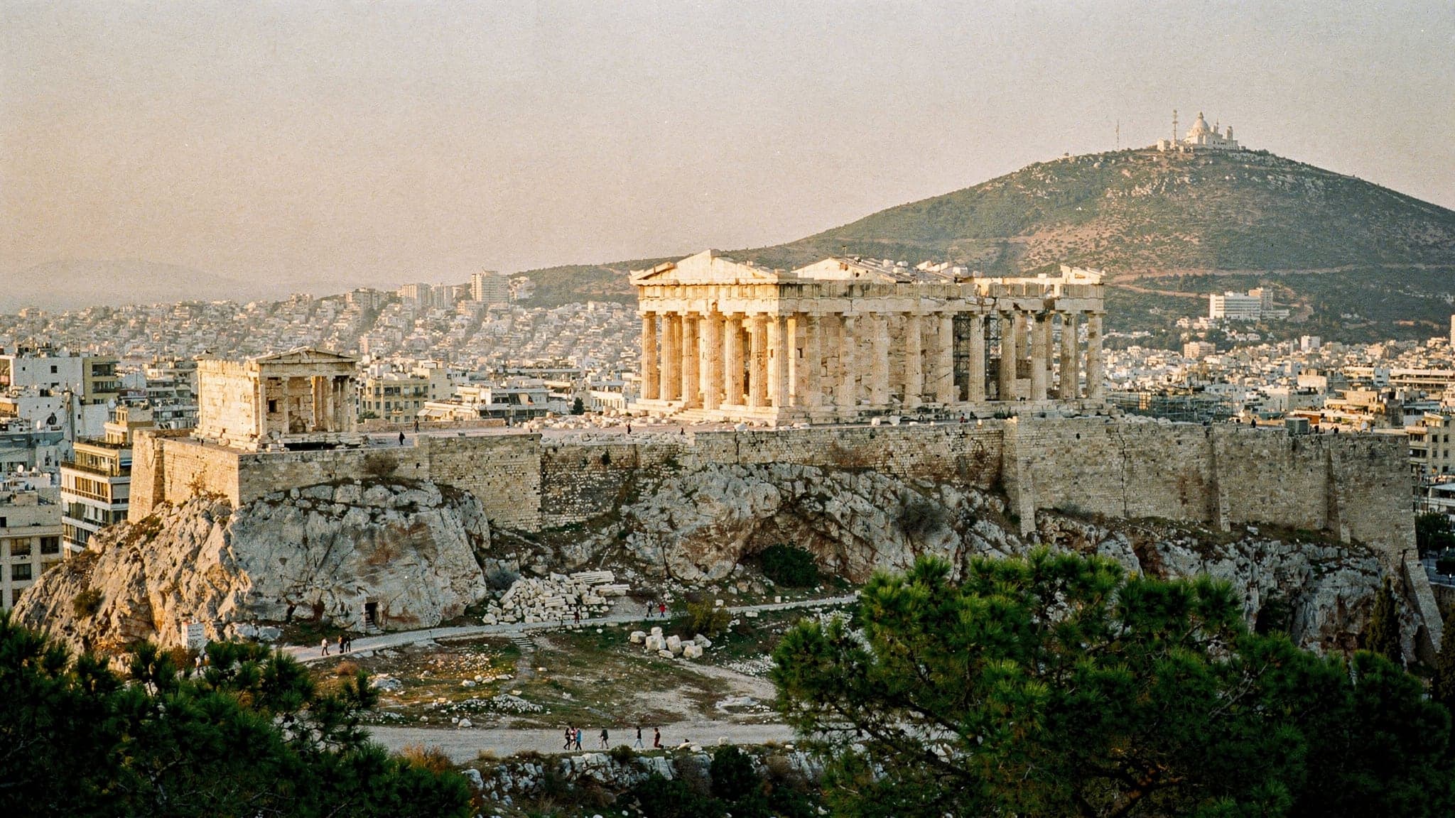 The Acropolis and the Parthenon at golden hour seen from Filopappou Hill, with the city's white apartment blocks spreading towards Mount Lycabettus