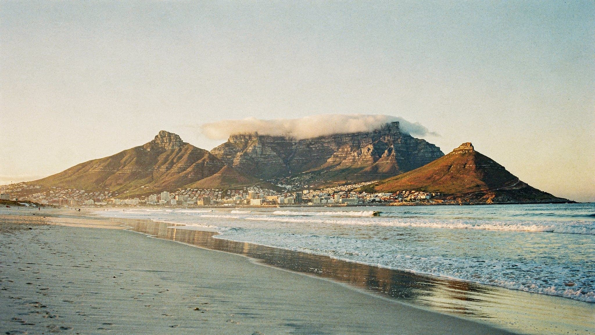 Table Mountain across Table Bay at golden hour, with a thin cloud spilling over its flat summit and waves breaking on the beach at Bloubergstrand