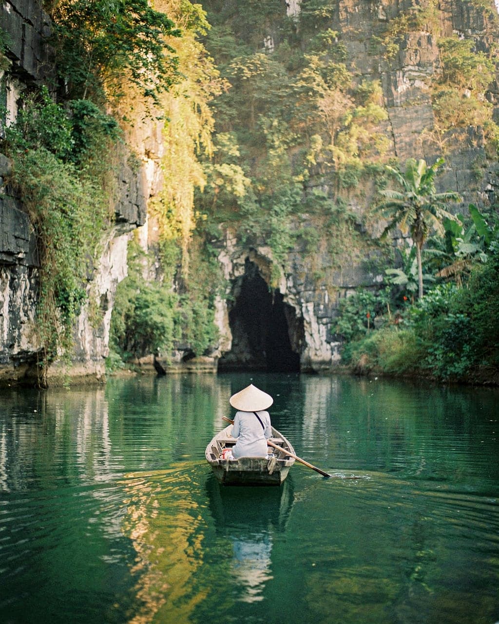 A rowing boat gliding between sheer limestone cliffs towards a low cave mouth at Trang An, Ninh Binh