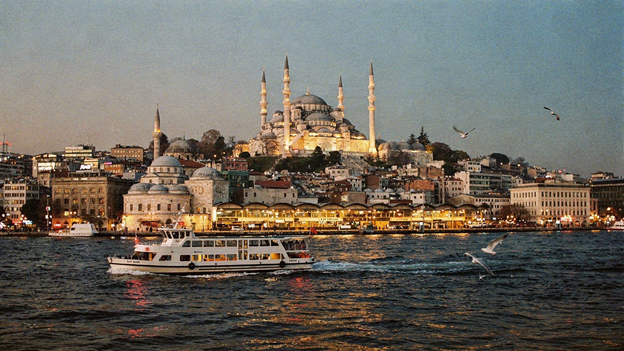 The Süleymaniye Mosque and the domes of the historic peninsula above the Golden Horn at dusk, with a commuter ferry crossing in the foreground