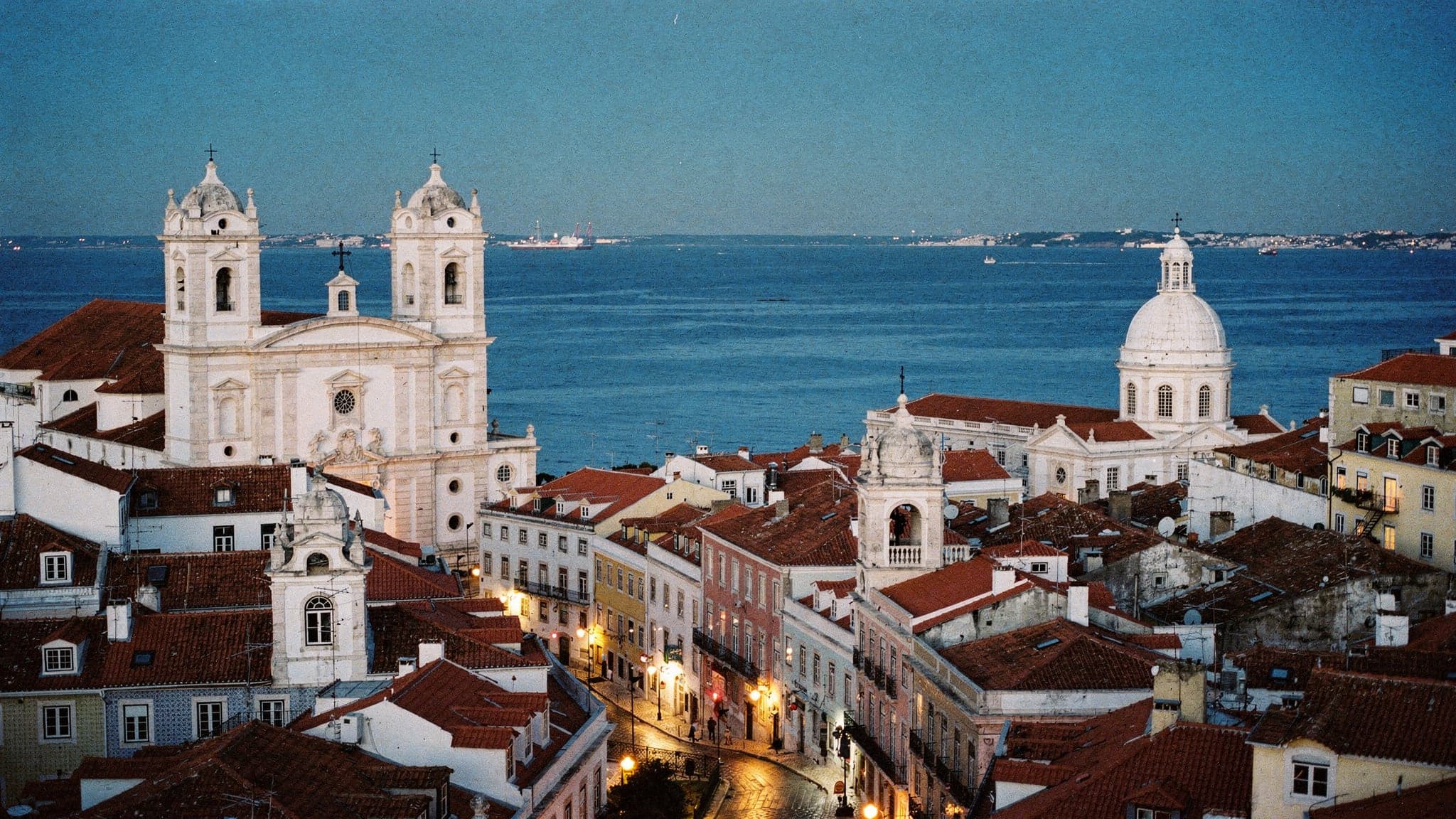 Alfama's terracotta rooftops and white church domes stepping down to the Tagus at blue hour, seen from the Miradouro das Portas do Sol