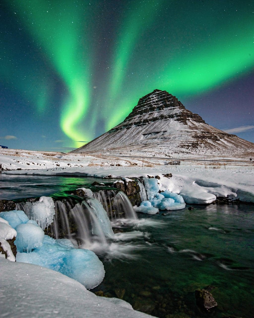 Green northern lights over Kirkjufell mountain and the frozen Kirkjufellsfoss waterfall on a winter night