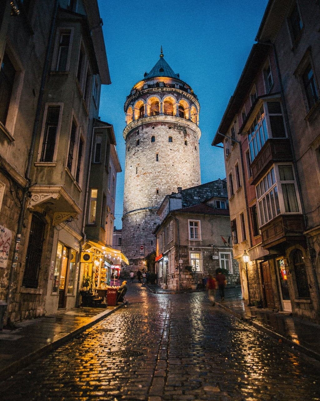 Galata Tower rising above a steep cobbled lane in Galata at blue hour, with lit shopfronts