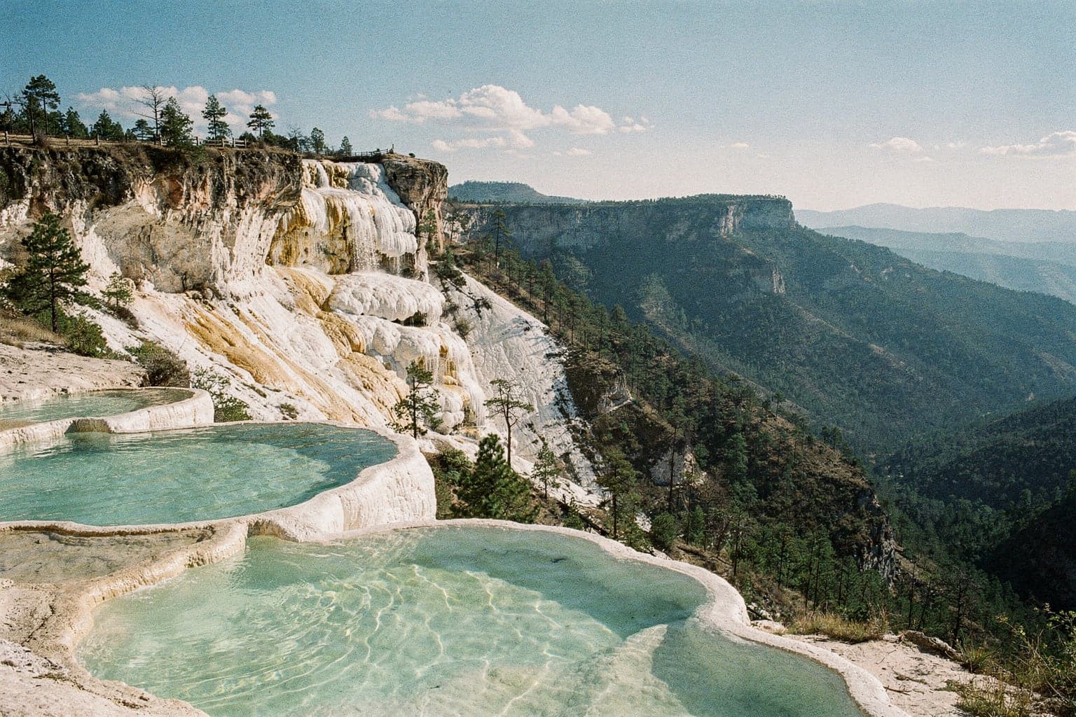 The petrified mineral waterfall of Hierve el Agua spilling down a cliff, with turquoise spring-fed pools at the edge overlooking forested ridges